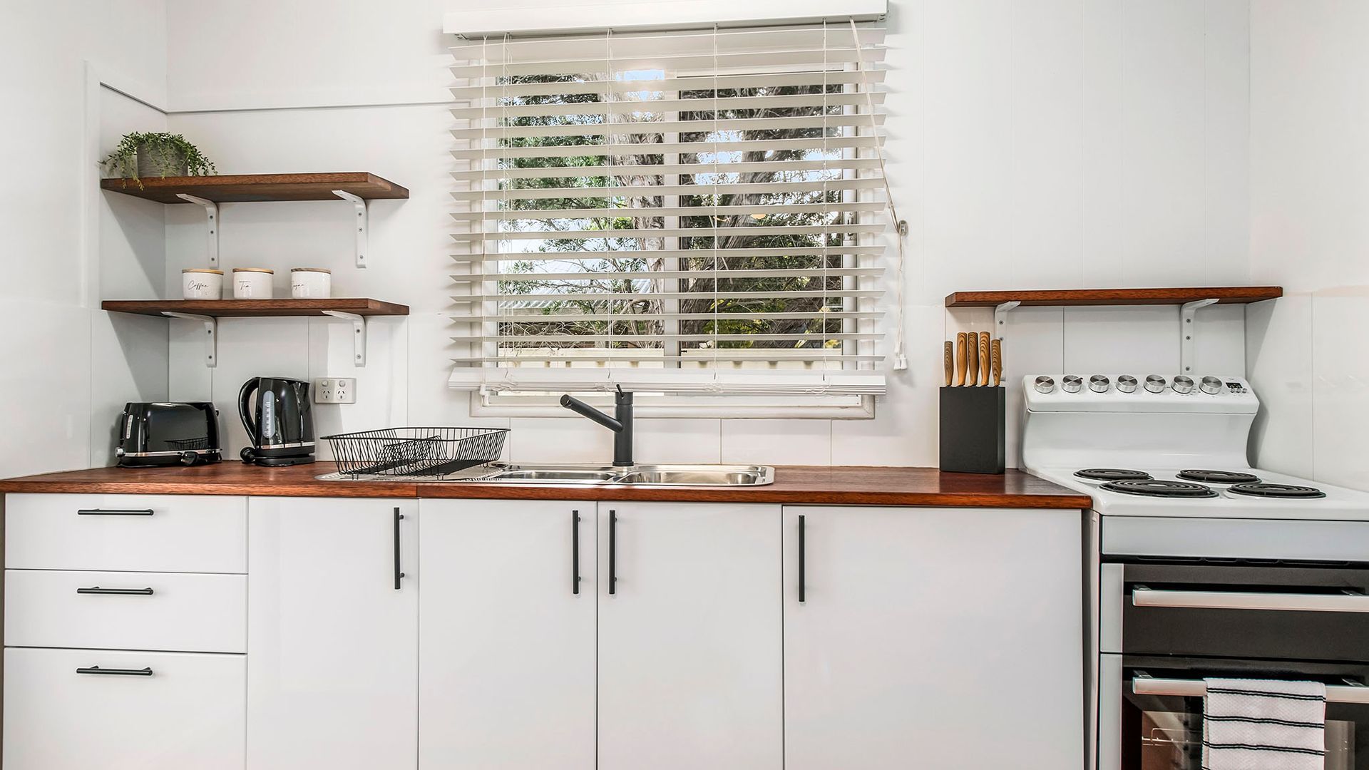 A modern kitchen with white cabinets, a wooden countertop, a built-in stove, and a window with blinds.