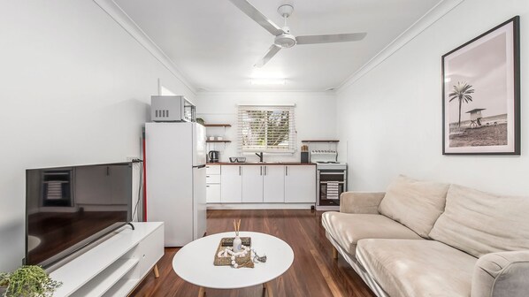 A modern living room with a flat-screen TV, a beige sofa, a white coffee table, and a kitchen area with white cabinets and a built-in oven.
