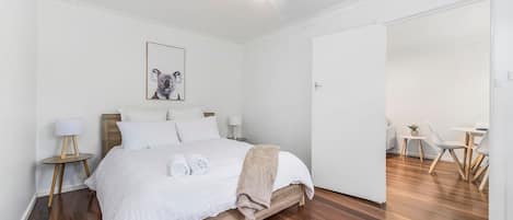 A hotel room with a wooden bed, white bedding, a koala artwork, a small table with a lamp, and a dining area with chairs.