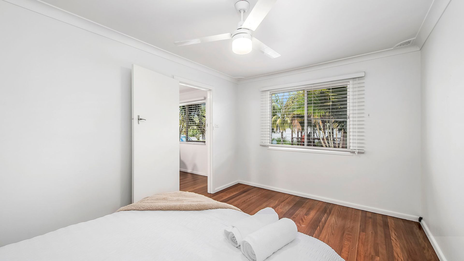 Quarto com cama, ventoinha no teto, janela com persianas e porta para outro cômodo.