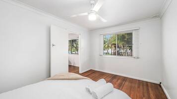 Quarto com cama, ventoinha no teto, janela com persianas e porta para outro cômodo.