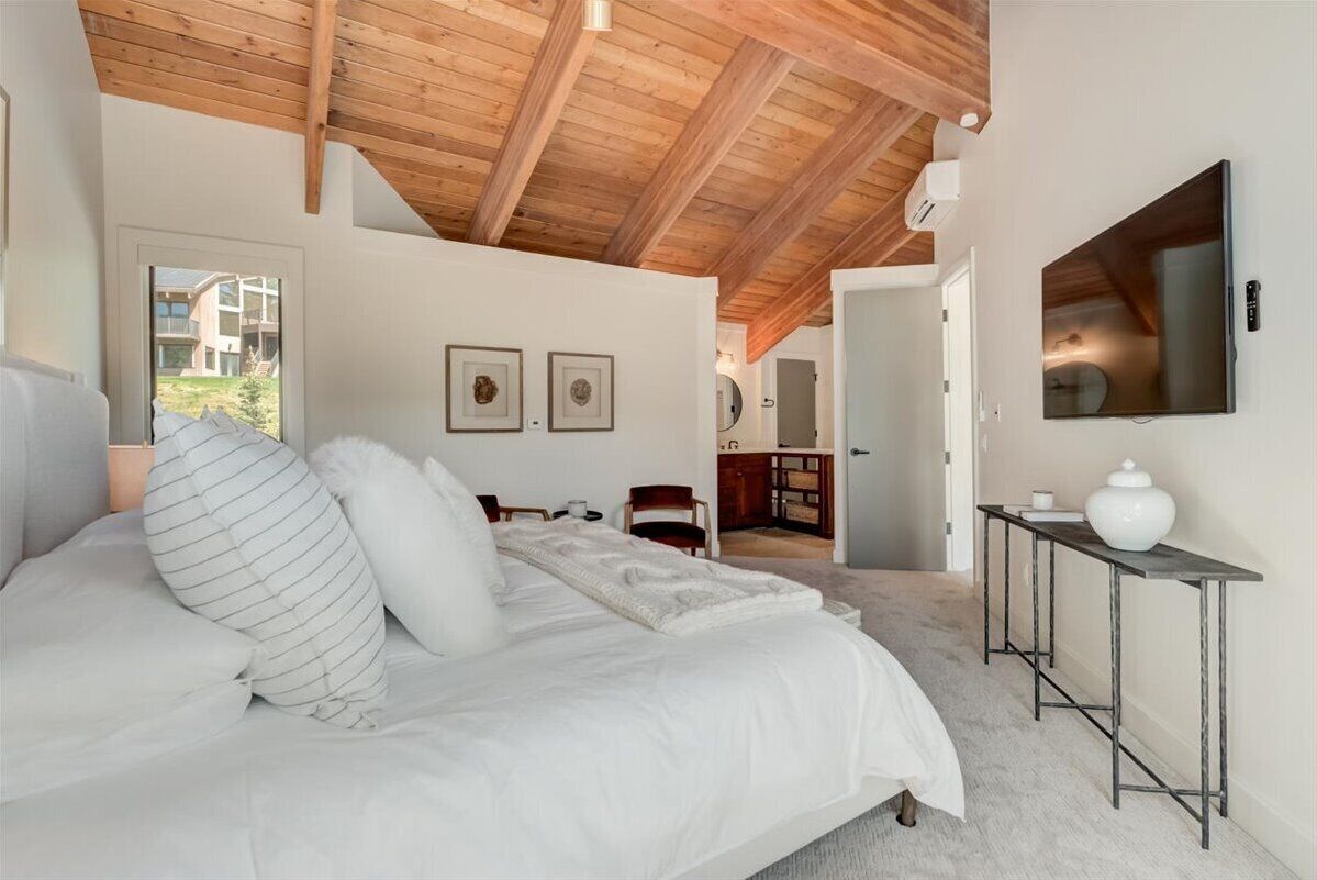 A bedroom with a large bed, wooden ceiling beams, a TV mounted on the wall, and a small table with a vase.