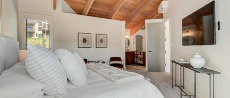 A bedroom with a large bed, wooden ceiling beams, a TV mounted on the wall, and a small table with a vase.