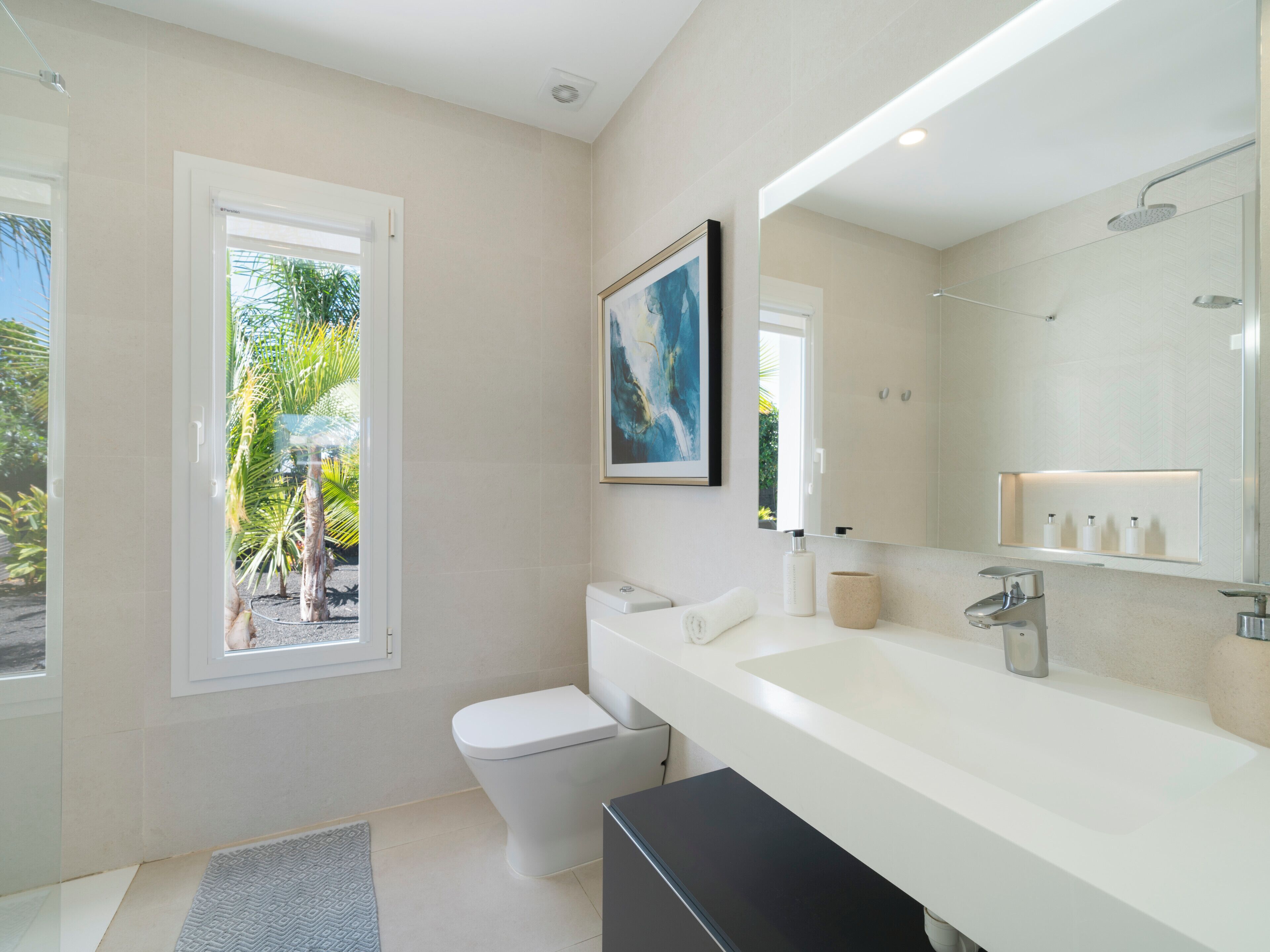 A modern bathroom with a toilet, sink, and shower. There is a window with a view of palm trees outside.