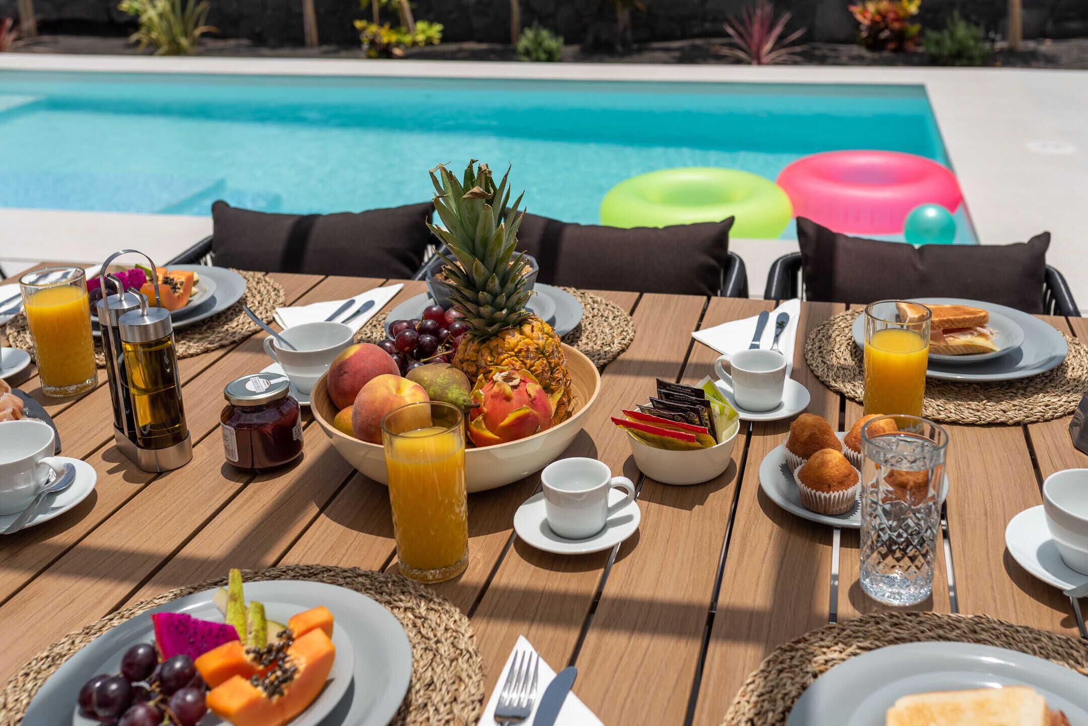A dining table set with breakfast items, including fruits, pastries, and beverages, by a poolside.