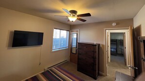 Una habitación con ventilador de techo, televisor montado en la pared, ventana con persianas, cómoda de madera y puerta que da a otra habitación.
