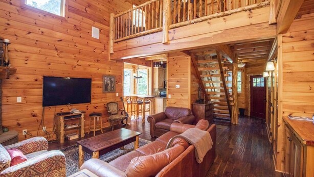 A spacious wooden cabin with a loft bedroom, a living room with leather sofas, a flat-screen TV, and a staircase leading to the upper level.