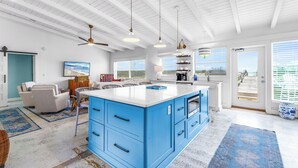 Una cocina amplia con una isla grande, vigas de techo blancas y vistas a la playa a través de puertas correderas de vidrio.