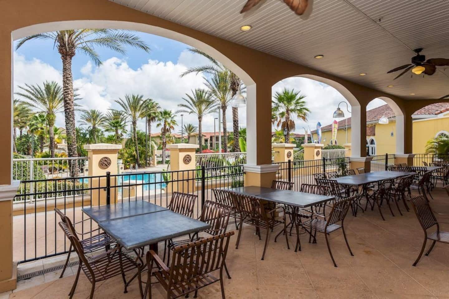 Un patio al aire libre con mesas y sillas, una piscina y palmeras al fondo.