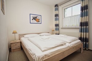 Un lit double bien fait, avec du linge de lit blanc, une table de chevet avec une lampe, un tableau encadré au mur et une fenêtre avec des rideaux.