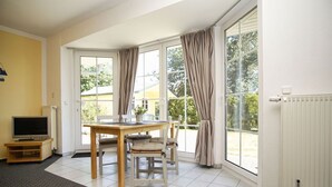 Une pièce avec une table et des chaises en bois, un téléviseur posé sur un meuble, et de grandes fenêtres avec des rideaux.
