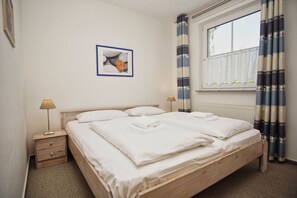 Un lit double bien fait, avec du linge de lit blanc, une table de chevet avec une lampe, un tableau encadré au mur et une fenêtre avec des rideaux.
