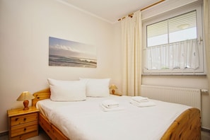 A bedroom with a wooden bed, white bedding, a nightstand with a lamp, a window with blinds, and a painting on the wall.