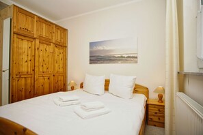 Een slaapkamer met een houten garderobe, een bed met witte lakens, twee bedlampen en een schilderij van een strandgezicht.