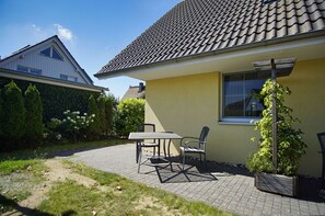 Uma casa com telhado em telhas, um pátio pavimentado com mesa e cadeiras, e um jardim com arbustos e uma árvore.