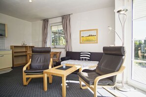 Un salon comprenant un canapé, deux fauteuils, une table basse et une table à manger. Il y a une fenêtre avec des rideaux, un tableau au mur et un coin cuisine avec des placards et un évier.