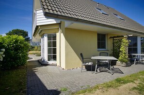 Une maison jaune avec un toit en tuiles, une terrasse avec une table et des chaises, et un jardin.