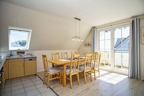 Un coin repas avec une table et des chaises en bois, une cuisine avec une fenêtre, et une vue sur un immeuble par le biais d’une grande fenêtre.