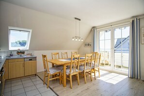 A dining area with a wooden table and chairs, a kitchen with a window, and a view of a building through a large window.