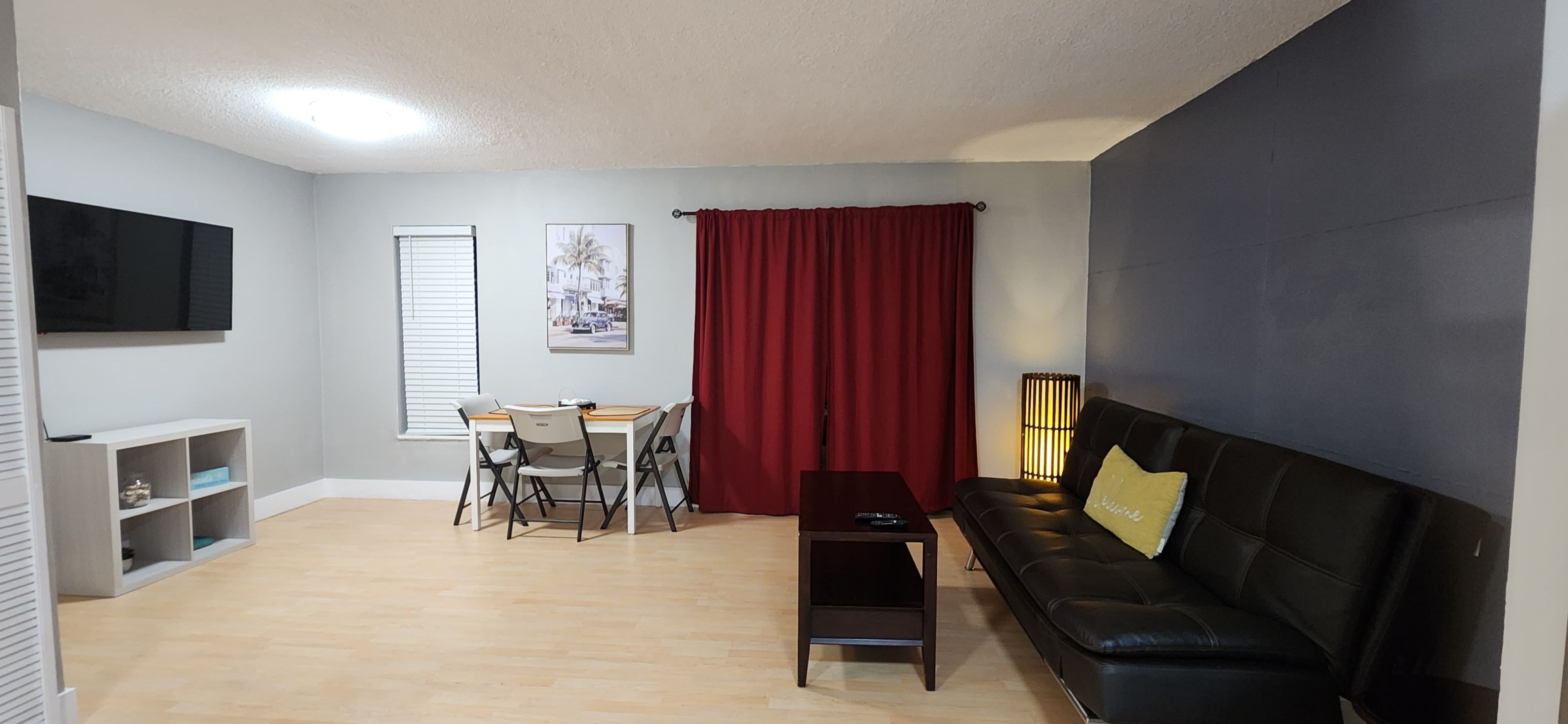 A living room with a black sofa, a dining table with chairs, a TV mounted on the wall, and a red curtain.