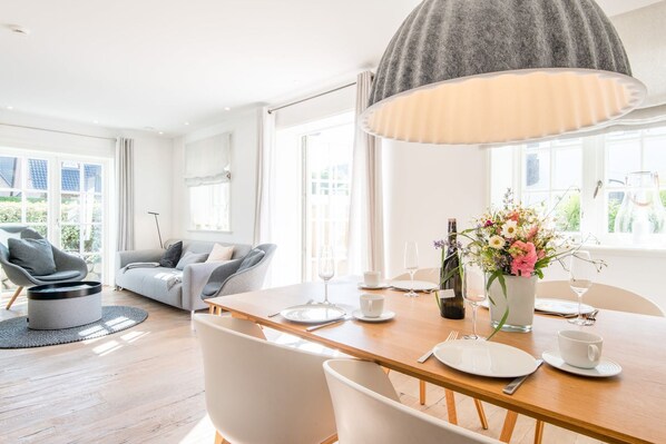 A modern living room with a dining area, featuring a round coffee table, a sofa, and a dining table set for two with wine glasses and a vase of flowers.