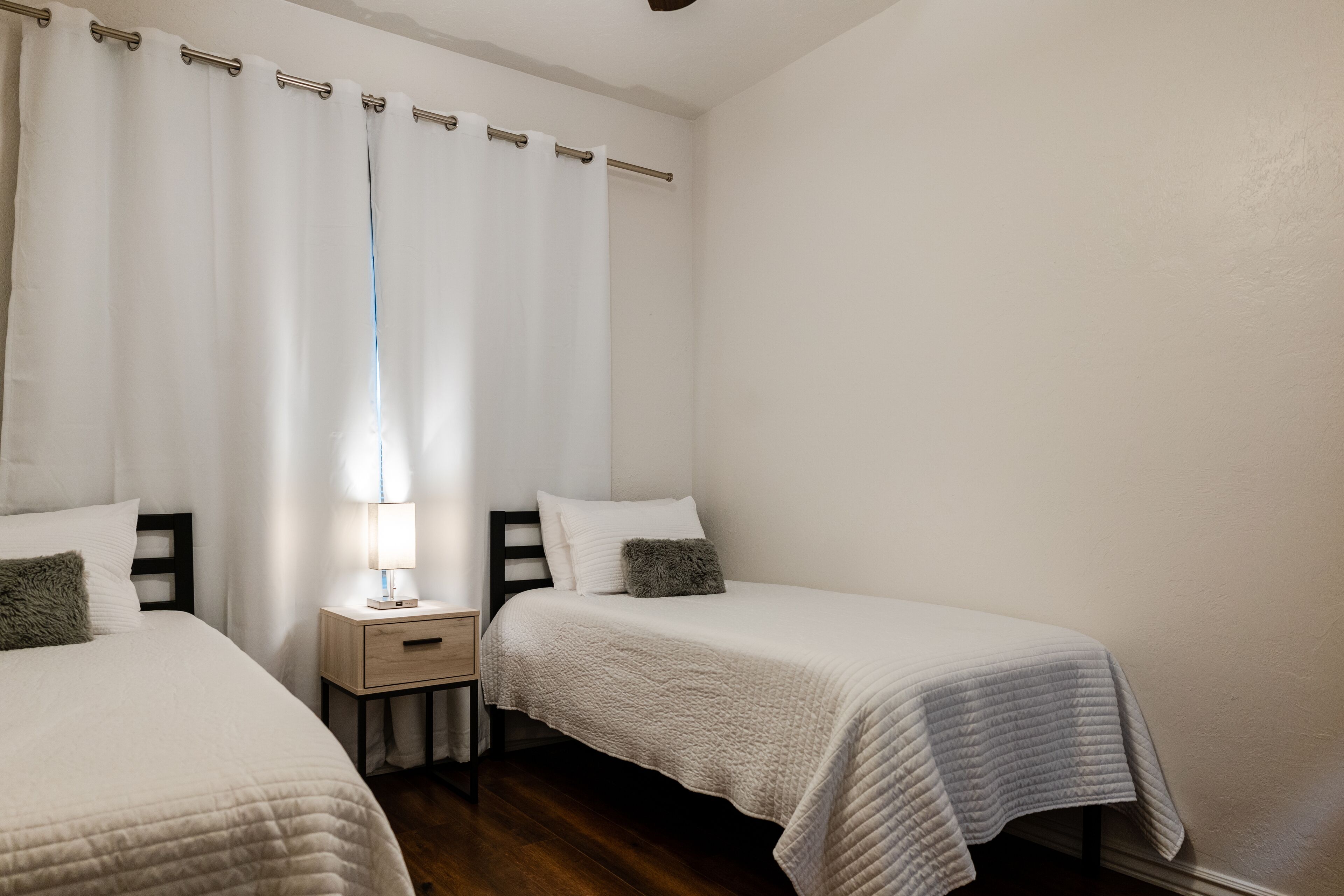 A bedroom with two beds, white bedding, a nightstand with a lamp, and a window with sheer curtains.