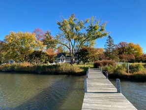 A wooden dock leading to a house by a calm body of water, surrounded by trees with autumn foliage.