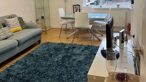 A living room with a grey sofa, a blue shag rug, a dining table with chairs, a ceiling fan, and a window with a dark blind. - West End 1-Bed Apt, Sleeps 2 with Wi-Fi (London)