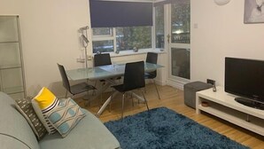 A modern living room with a glass dining table, a grey sofa, a blue rug, and a TV on a white stand. - West End 1-Bed Apt, Sleeps 2 with Wi-Fi (London)