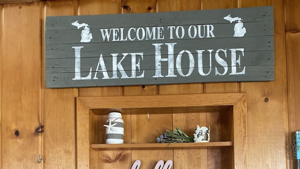 Un mur en bois orné d’une pancarte « Bienvenue dans notre maison au bord du lac », une étagère avec des objets décoratifs et une pancarte « Bonjour ».