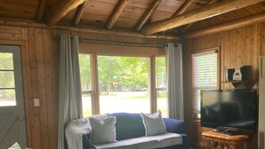 Une pièce chaleureuse avec un plafond en bois, un canapé, un téléviseur posé sur un meuble en bois et une grande fenêtre avec des rideaux.