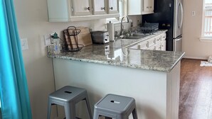 A kitchen with white cabinets, a black refrigerator, a granite countertop, and two grey stools. - Sargent Beach House Getaway - fish from the dock and walk to the beach! (Sargent)