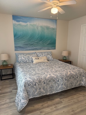 A bedroom with a bed, two nightstands, a ceiling fan, a wall painting of a wave, and a door. - Sargent Beach House Getaway - fish from the dock and walk to the beach! (Sargent)