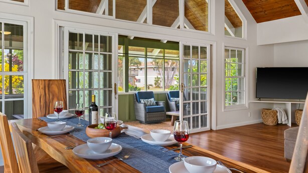 A dining area with a long wooden table set for four, surrounded by large windows and a view of the outdoors.