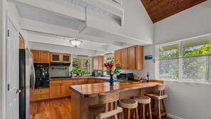 A kitchen with wooden cabinets, a central island with stools, and a view of greenery through the window.
