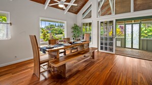 A spacious dining area with a wooden table and benches, large windows with views of greenery, and a ceiling fan.