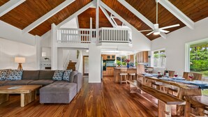 A spacious living area with a high ceiling, wooden flooring, a dining area with wooden tables, and a kitchen in the background.