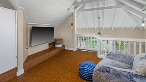 Ein geräumiges Dachzimmer mit Holzboden, weißer Decke mit sichtbaren Balken, weißer Geländer und einem großen Fenster mit Blick auf Grünflächen.