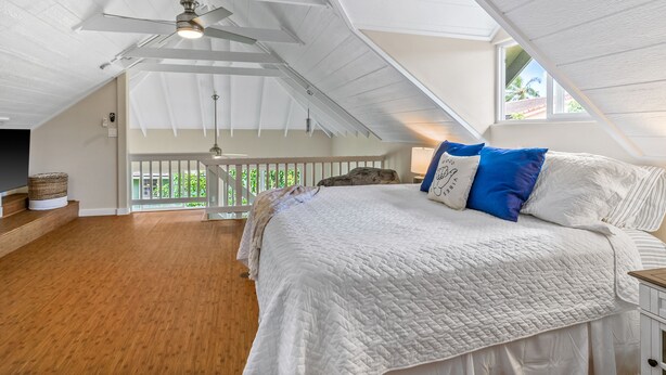 Ein Schlafzimmer mit schrägen Wänden, einem Bett mit weißer Bettwäsche und blauen Kissen, einem Holzboden und einem Balkon mit Geländer.