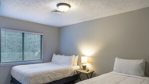 Ein Schlafzimmer mit zwei Betten, einem Fenster mit Jalousien, einem Nachttisch mit Lampe und einer Deckenleuchte.