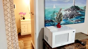Una cocina con un microondas sobre una mesa de madera, un cuadro de un faro y un baño con cortina de ducha.