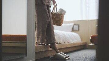 Una persona con bata entrando a una habitación con una cama, un banco de madera y una cesta en el suelo.