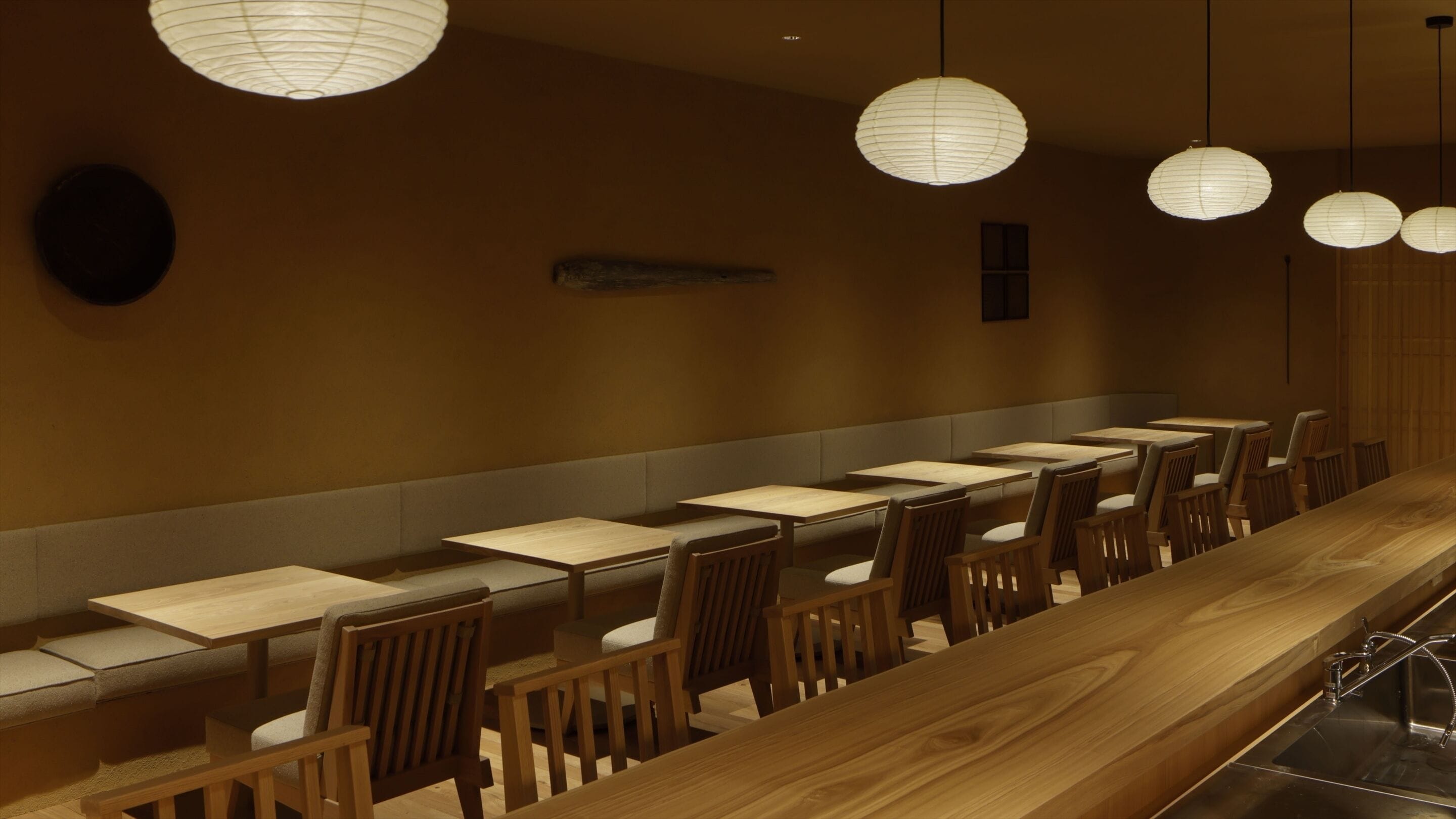 A restaurant with wooden tables and chairs, paper lanterns hanging from the ceiling, and a long counter with a sink.