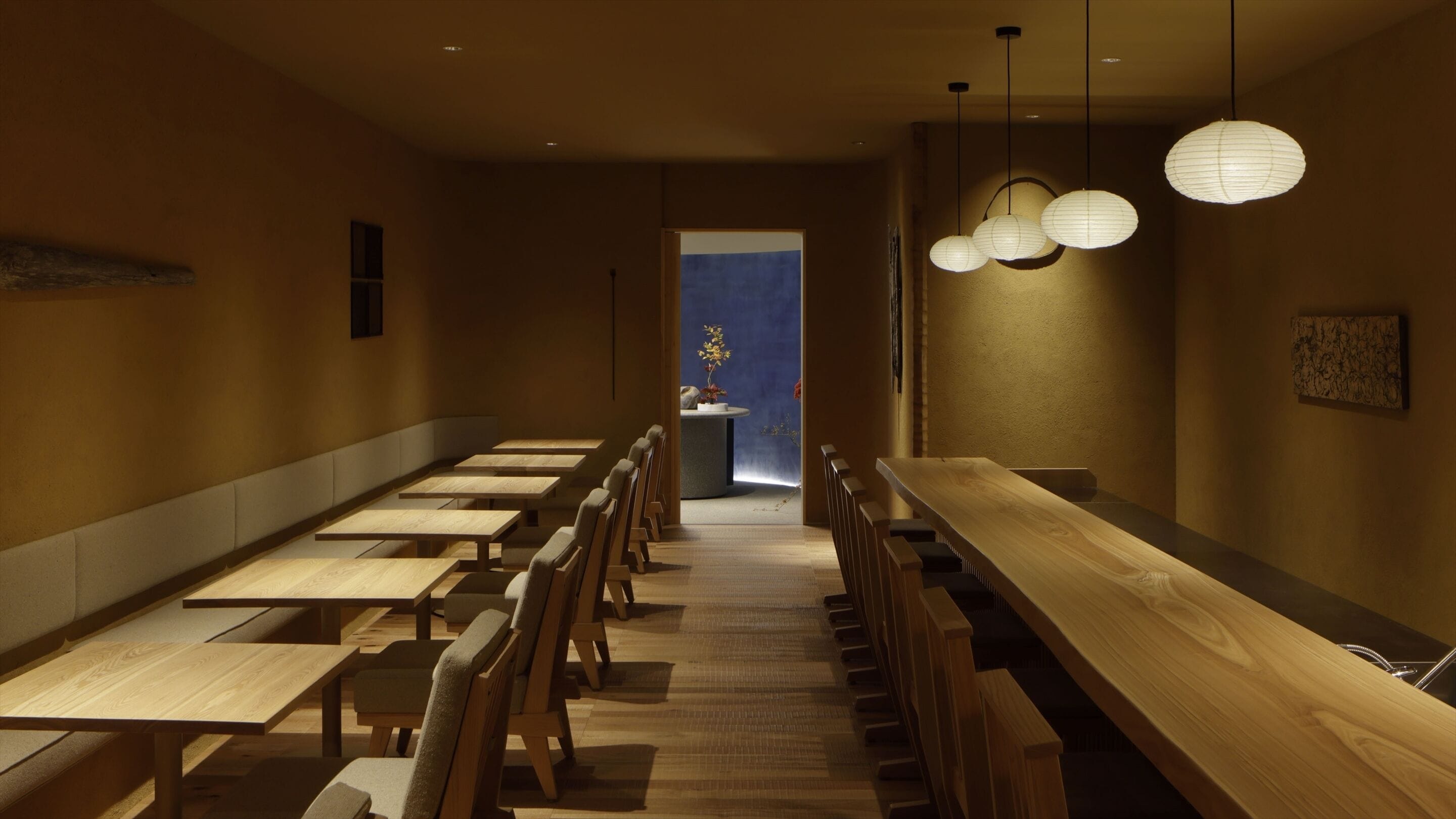 A restaurant with long wooden tables and chairs, a counter with a blue backdrop, and hanging paper lanterns.