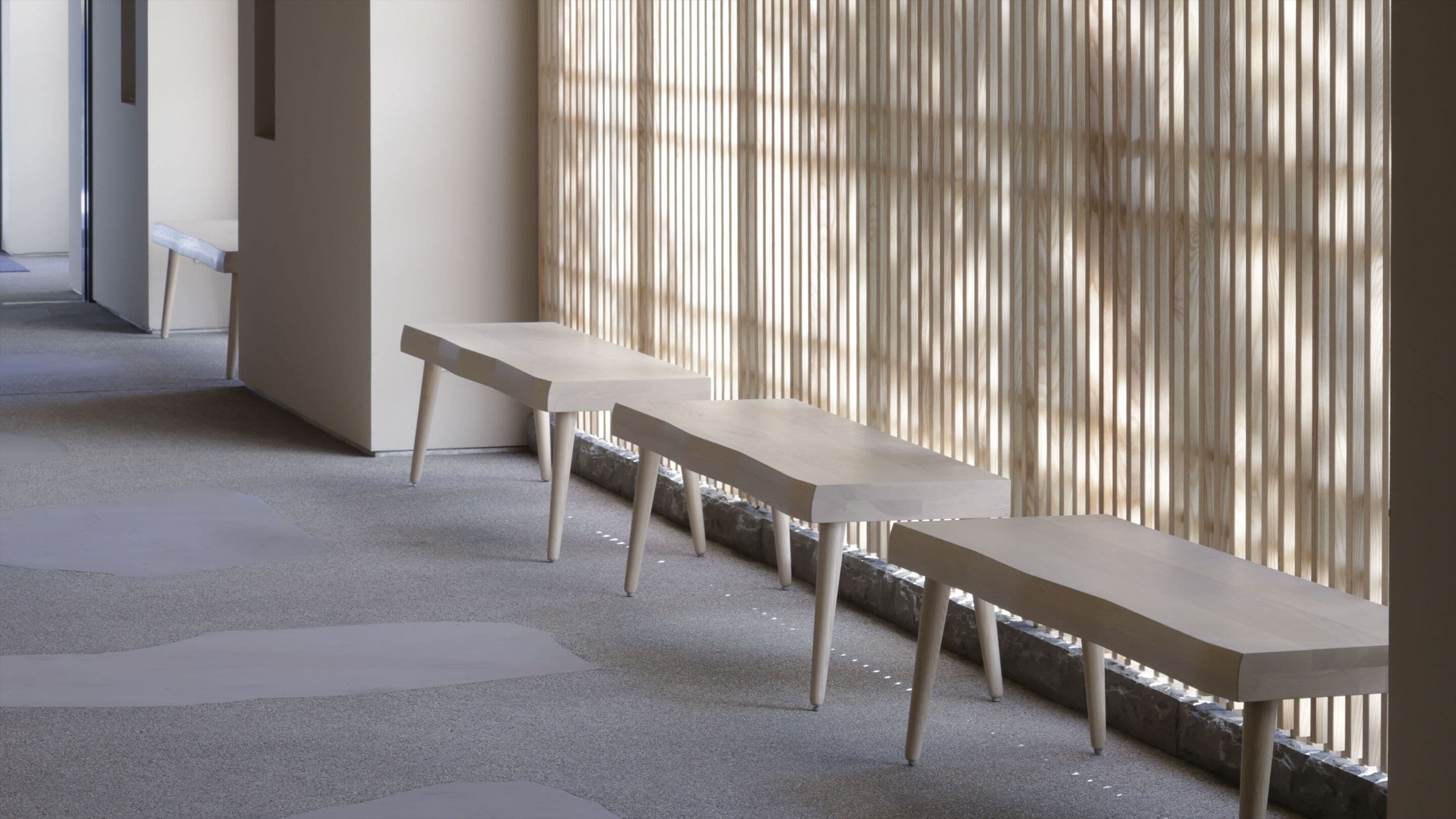 Three minimalist wooden benches aligned against a wall with vertical blinds, in a corridor with a speckled floor.