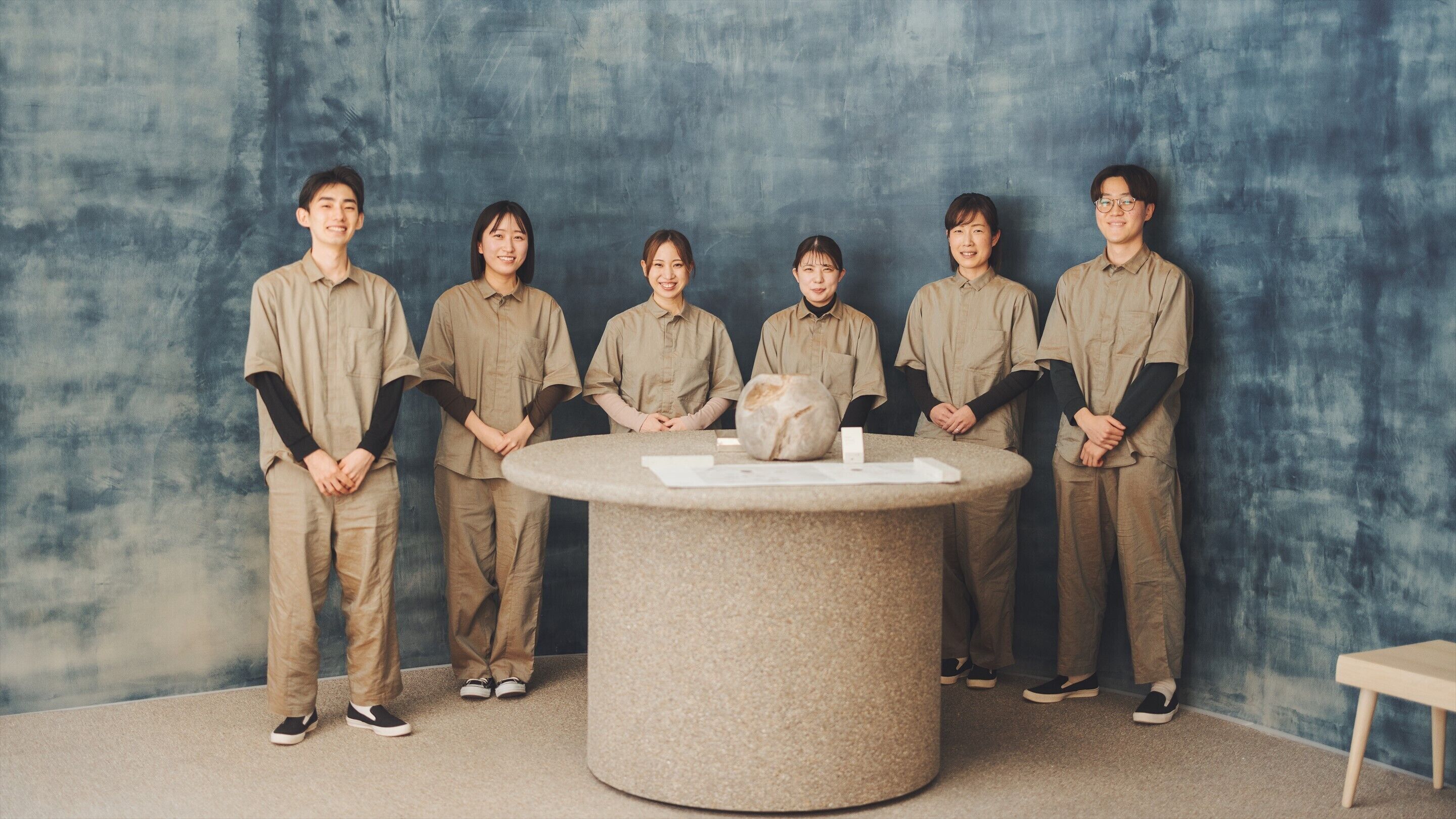 A group of six people in uniform standing around a round table with a stone top, against a textured blue wall.