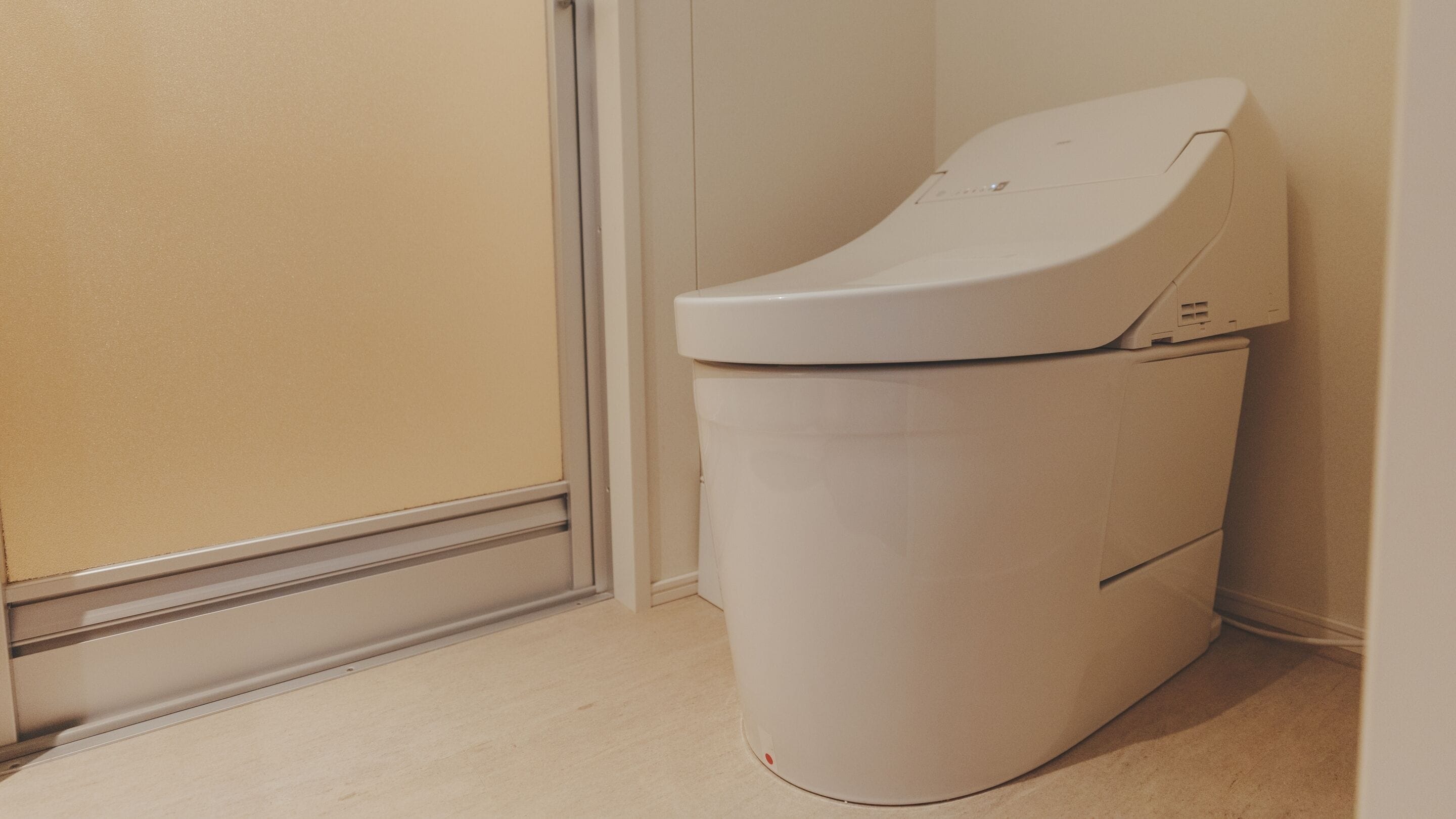 A compact, white, wall-mounted toilet in a narrow hallway with a beige floor and walls.