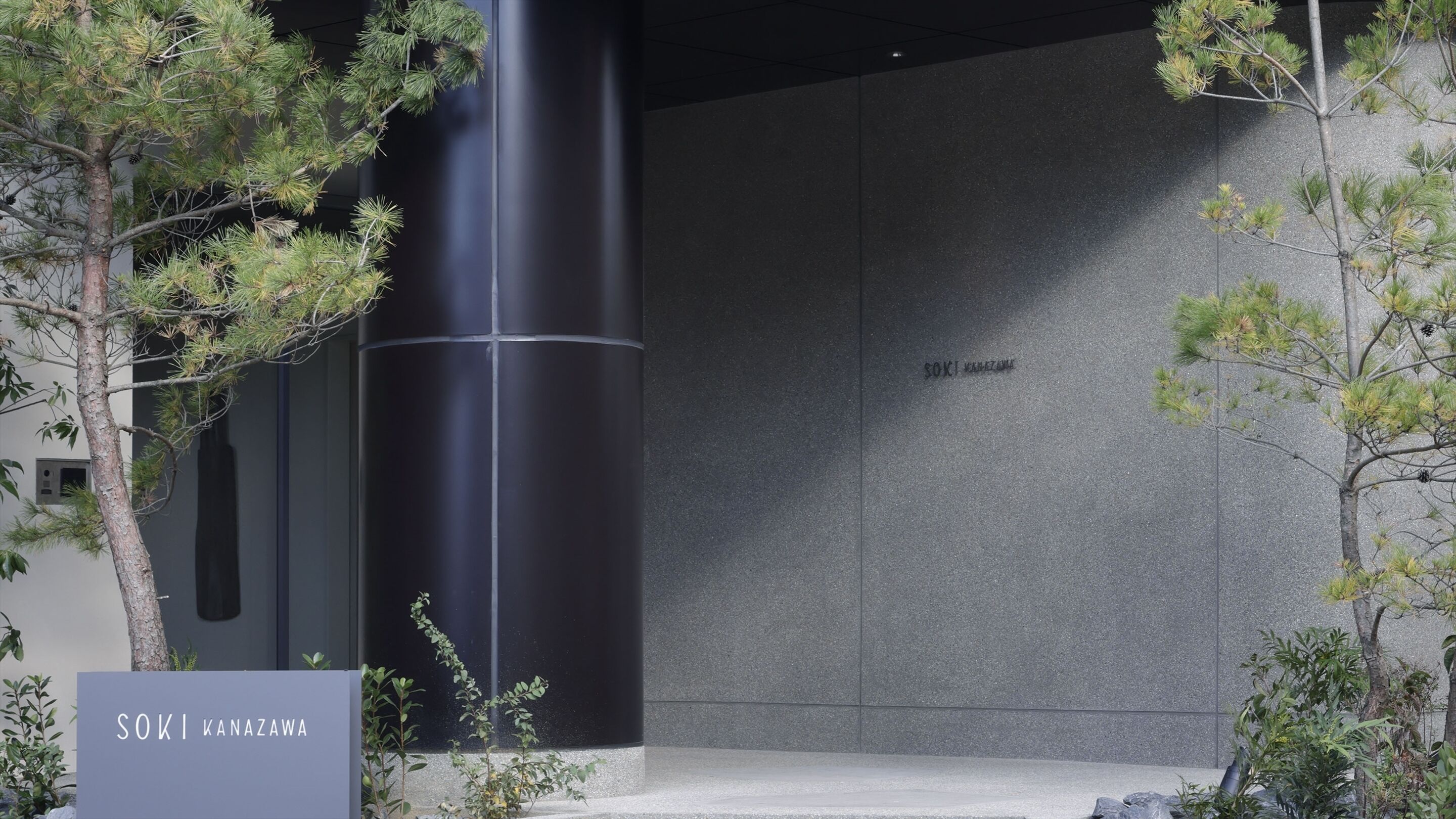 A modern building entrance with a tall cylindrical structure, a nameplate reading "SOKI KANAZAWA", and surrounding greenery.