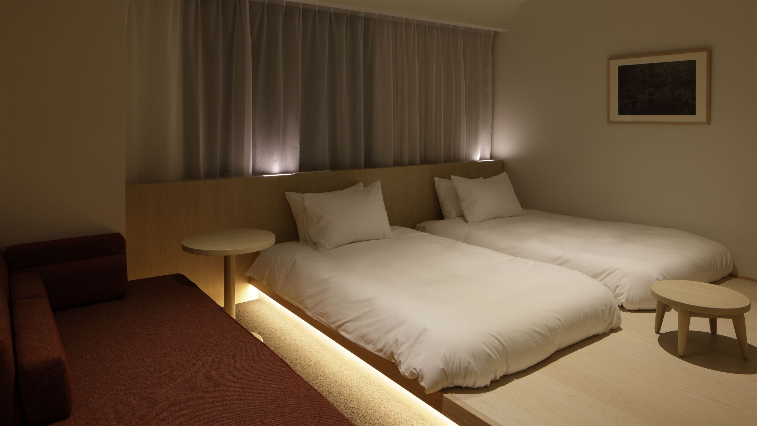 A hotel room with two single beds, a small table, a bench, and a painting on the wall.