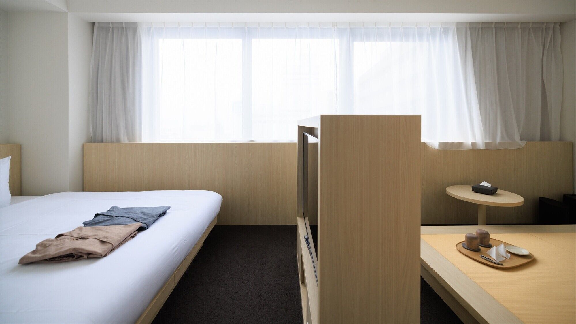 Une chambre d’hôtel avec un lit, un bureau sur lequel se trouve un plateau avec divers objets, et une fenêtre avec des rideaux.